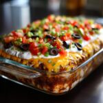 A close-up view of a freshly made 7-layer dip in a glass baking dish, topped with tomatoes, olives, and green onions.