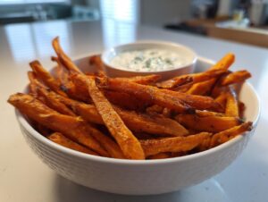 A white bowl filled with crispy air fryer sweet potato fries and a small dish of dipping sauce.