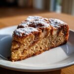 A slice of moist apple cake dusted with powdered sugar, showcasing tender apple pieces within.