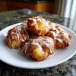 A close-up of several golden brown apple fritters drizzled with a white glaze on a white plate.