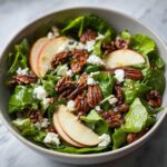 A close-up of a fresh Apple Pecan Salad in a bowl, featuring spinach, apple slices, pecans, and crumbled feta cheese.