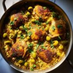 A close-up overhead view of a pan filled with flavorful Arroz con Pollo, featuring golden-brown chicken thighs, yellow rice, green olives, and fresh parsley.