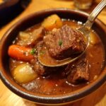 A spoonful of rich beef stew, showcasing tender chunks of beef, carrots, and potatoes in a savory broth.