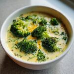 A close-up of a white bowl filled with creamy broccoli cheddar soup, featuring tender broccoli florets and melted cheddar cheese.