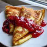Close-up of golden-brown cheese blintzes folded and topped with vibrant raspberry sauce.