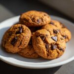 A pile of freshly baked chewy pumpkin chocolate chip cookies on a white plate, with visible chocolate chunks.