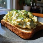 A close-up of a classic egg salad sandwich on toasted bread, topped with fresh dill.
