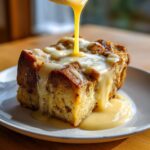 A slice of creamy bread pudding being drizzled with rich vanilla sauce.