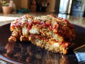 A close-up of a slice of crispy Eggplant Parmesan, showing layers of breaded eggplant, marinara sauce, and melted mozzarella cheese.