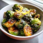 Close-up of crispy roasted Brussels sprouts topped with grated Parmesan cheese and lemon zest in a white bowl.