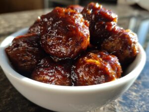 A close-up of a white bowl filled with glossy Crockpot BBQ meatballs, coated in a rich, dark sauce.