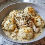 A close-up of a bowl of Crockpot chicken and dumplings, topped with shredded chicken and fresh herbs.
