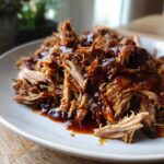 A close-up of tender Crockpot pulled pork generously coated in a rich BBQ sauce on a white plate.