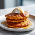 A stack of three fluffy pumpkin pancakes topped with whipped cream and drizzled with syrup.