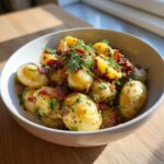 A close-up of a bowl of warm German potato salad, featuring small boiled potatoes, crispy bacon bits, and fresh dill.