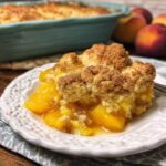 A close-up of a serving of Grandma's Peach Cobbler on a white plate, with a fork and fresh peaches in the background.