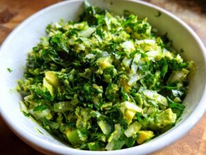 A close-up, overhead view of a bowl filled with vibrant Green Goddess Salad, featuring chopped avocado, fresh herbs, and onions.