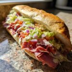 A close-up of a loaded Grinder Salad Sandwich with deli meats, lettuce, tomatoes, red onion, and creamy dressing on a baguette.