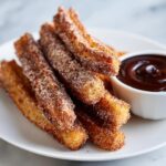 A close-up of a pile of freshly made homemade churros, coated in cinnamon sugar, served with a small bowl of chocolate sauce.