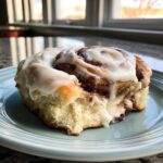 A close-up of a fluffy homemade cinnamon roll generously topped with creamy white frosting.