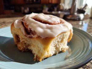 A close-up of a fluffy homemade cinnamon roll topped with creamy white frosting, on a blue plate.