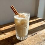 A tall glass of creamy Horchata filled with ice and topped with cinnamon powder and two cinnamon sticks.