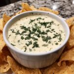 A white bowl filled with creamy Queso Blanco Dip, garnished with chopped cilantro, surrounded by tortilla chips.