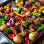 A close-up view of a sheet pan filled with roasted sausage and colorful vegetables including broccoli, bell peppers, red onion, and potatoes.