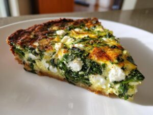 A close-up of a slice of spinach feta quiche on a white plate, showcasing the creamy filling and golden crust.