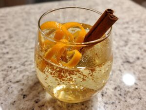 A close-up of an Apple Cider Spritz Cocktail in a glass, garnished with orange peel and a cinnamon stick.