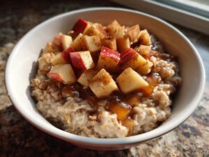 A close-up of a white bowl filled with warm Apple Cinnamon Oatmeal, topped with diced apples and a drizzle of syrup.