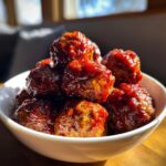 A close-up shot of a white bowl filled with glistening BBQ Meatballs, coated in a rich, savory sauce.