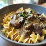 A close-up of a bowl of Beef Stroganoff served over wide egg noodles, garnished with fresh parsley.