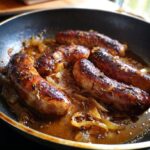 Close-up of Beer Braised Bratwurst sizzling in a pan with caramelized onions and sauce.