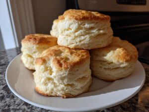 A stack of golden-brown, flaky buttermilk biscuits on a white plate, showcasing their layered texture.