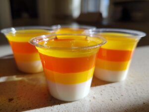 Close-up of layered Candy Corn Jello Shots in clear plastic cups, showcasing white, orange, and yellow layers.