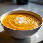 A close-up of a white bowl filled with vibrant orange Carrot Ginger Soup, topped with a swirl of cream.