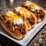 Three loaded chili dogs topped with chili, shredded cheese, sour cream, and diced red onion.