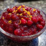 A close-up of vibrant, homemade Cranberry Sauce in a glass bowl, topped with fresh orange zest.