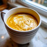 A close-up of a white mug filled with creamy Pumpkin Soup, swirled with cream and black pepper.