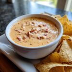 A bowl of creamy Queso Dip with red pepper pieces and herbs, surrounded by tortilla chips.