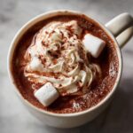 A close-up of a mug filled with rich, decadent hot chocolate, topped with whipped cream, marshmallows, and a dusting of cocoa powder.
