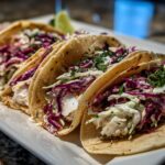 Close-up of three freshly made Fish Tacos filled with flaky white fish, vibrant red cabbage slaw, and fresh cilantro, served with lime wedges.