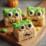 Three Frankenstein Rice Krispie Treats decorated with green frosting hair and candy eyes, sitting on a wooden board.
