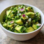 Close-up of a bowl of fresh guacamole, featuring chunky avocado pieces, diced red onion, and cilantro.