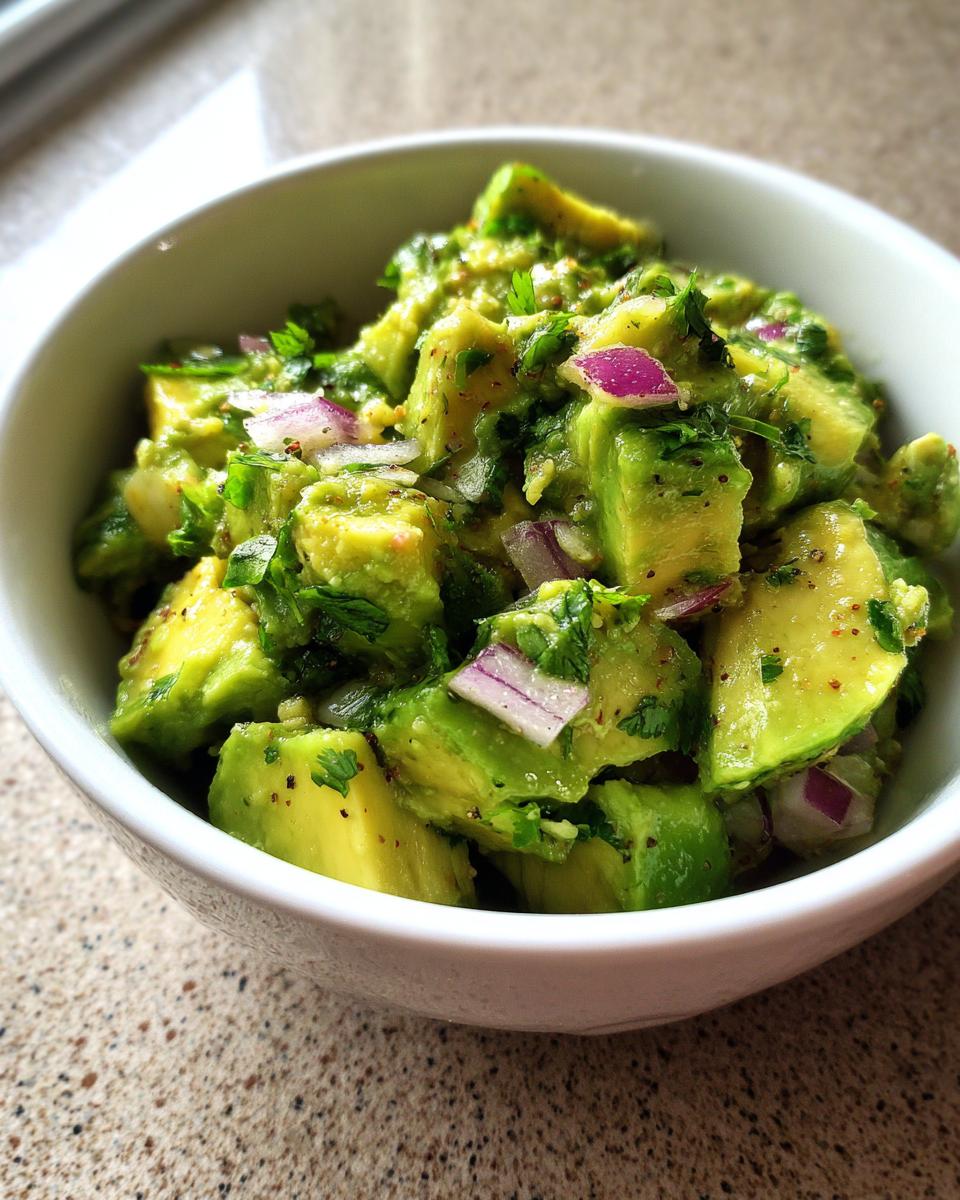 A close-up of chunky guacamole in a white bowl, featuring ripe avocado chunks, red onion, and cilantro.