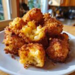 A close-up of a stack of golden-brown Fried Mac and Cheese Bites on a white plate, with one bite broken open to reveal gooey cheese.