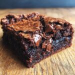 A close-up of a rich, fudgy brownie with melted chocolate chips on a wooden surface.