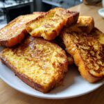A close-up of golden brown French toast slices, glistening with syrup, piled on a white plate.