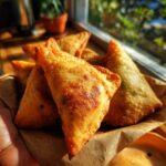 A close-up shot of golden-brown, crispy samosas piled in a brown paper basket, ready to be served.